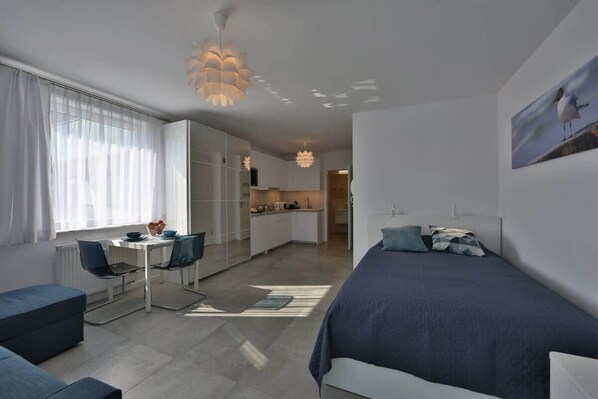 A stylish studio living area with a cozy sofa, a small dining table, and large windows that allow plenty of natural light, giving a bright and open feel.