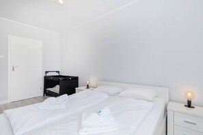  Simple bedroom with a bed, white linens, and a small desk area. A window offers some natural light.