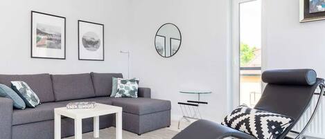 Bright living room with a gray sofa, a coffee table, and a wall-mounted TV. Large windows let in plenty of natural light.