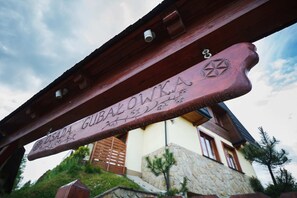 Close-up view of a wooden building sign under a sloped roof, with a portion of the building’s exterior visible.