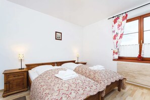  A simple bedroom with a single beds covered in floral bedding, a wooden nightstand with a lamp, and a small window offering natural light.