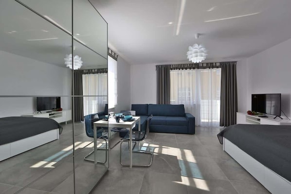 A modern living room featuring a sofa, dining table, and a chandelier, with bright natural light coming through large windows.