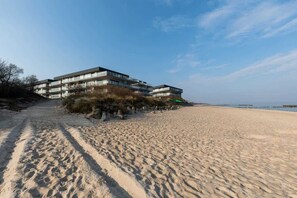 A beach view showing the sandy shore, with the apartment complex visible in the background and a clear, blue sky.