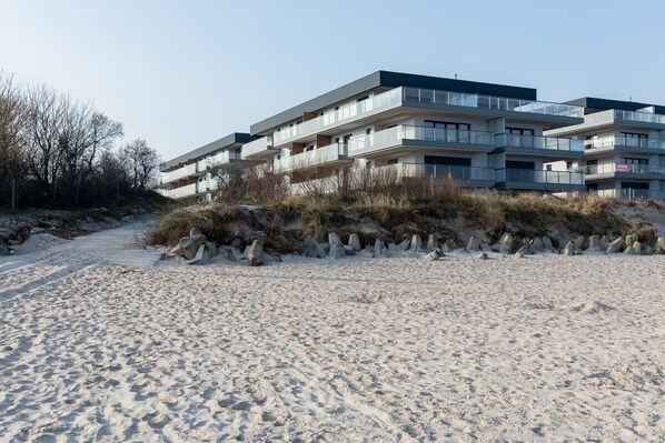 An exterior shot of a modern apartment complex with large balconies, situated near a sandy beach with a clear view of the building.