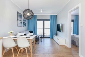 A bright and spacious living room featuring a dining table with white chairs, a grey sofa, and large glass doors leading to a balcony.