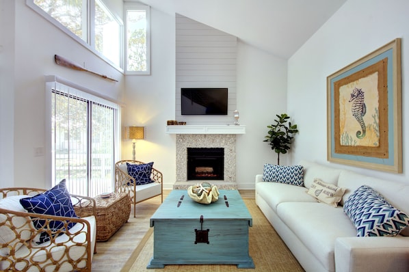 Vaulted Living Room • Stylish Coastal Design • St. Simons - Step into Sea Glass Cottage’s bright, beach-chic living room—complete with soaring ceilings, a gas fireplace, smart TV, and natural light that fills every corner. The coastal color palette, seahorse art, and warm wood accents create a welcoming space to relax, nap, or plan your next beach day.
