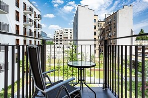 The image shows a residential balcony with outdoor furniture. In the background, there's a modern apartment complex surrounded by greenery and open spaces.