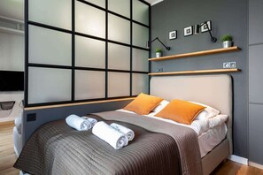 A bedroom with a double bed covered in neutral-colored bedding. The room features a large industrial-style window and minimalist décor.