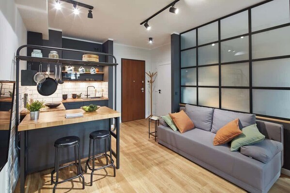 A cozy living room with a large gray couch, modern industrial lighting, and a small dining table. There’s a wooden floor, black window frames, and minimalist décor, giving a warm, open feel.