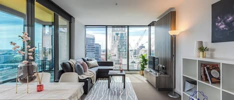 An open-plan space features a living area furnished with an L-sofa with a fold-out sofa bed, floor-to-ceiling windows, a wooden coffee table, and a wide-screen TV.