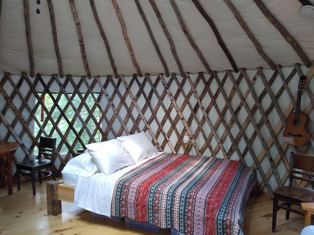 A bed in a Maine glamping yurt is seen with lattice work surrounding, a guitar hanging on the wall, and colorful bedding.