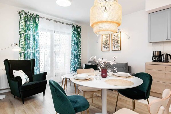 A cozy living room featuring a round dining table, green velvet chairs, and a large window with patterned green curtains. The space is bright and stylish.