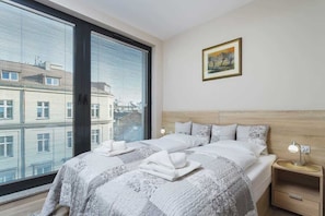 A bedroom with a large double bed, two side tables, and floor-to-ceiling windows providing an urban view. The room is decorated in a minimalist style with neutral tones.