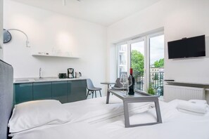 A bright living room with a sofa and TV, decorated in neutral tones, with glass doors leading to a balcony that brings in plenty of natural light.