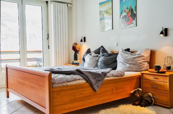 Bedroom with large bed, wardrobe, TV and large window front with access to the covered balcony overlooking St. Moritz.