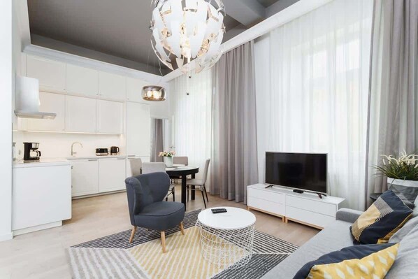 A cozy living room featuring a large sofa, a coffee table, and a TV. The room is well-lit with natural light coming from big windows.