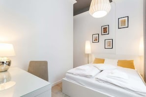  A neatly arranged bedroom with a double bed covered in white linens. The room has a nightstand with a lamp and decorative elements on the wall.