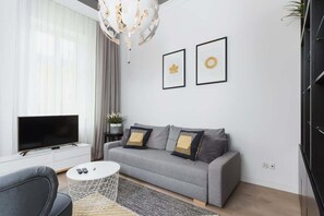 A living room view with a sofa, small round coffee table, and a flat-screen TV mounted on the wall. Light curtains allow natural light to brighten the room.