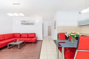 A spacious living room with a red corner sofa, coffee table, and a TV stand. The open-plan design connects to the kitchen in the background.