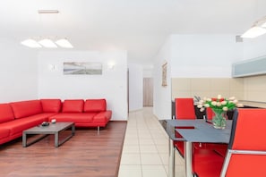 A spacious living room with a red corner sofa, coffee table, and a TV stand. The open-plan design connects to the kitchen in the background.