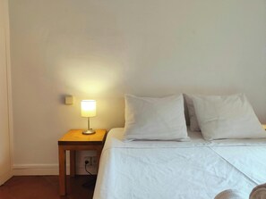 This inviting bedroom features a queen-size bed with crisp white linens and a bedside table with a lamp for a touch of cozy ambience. The white walls and simple décor create a calming atmosphere perfect for a restful night's sleep.