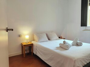 The bedroom features a queen-size bed with crisp white linens and a bedside table with a lamp, providing a cozy and inviting space for relaxation. The room is bathed in natural light streaming in from a window with black shutters. The terracotta tiled flo