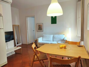 The living room is comfortably furnished with a white couch and a wooden dining table that seats four. The room is bright and airy, with tiled floors and a large window that lets in natural light. The apartment is located in a quiet area, but is close to 