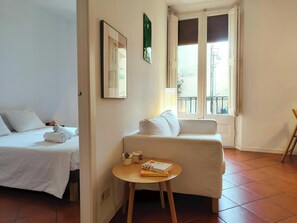 The spacious living area is bathed in natural light streaming through the French balcony doors. A comfortable white sofa sits invitingly near the window, perfect for relaxing after a day of exploring the city. The tiled floor adds a touch of classic Barce