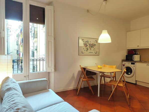 The apartment's living area features a white sofa placed in front of a window with white shutters. The window offers a view of a charming, narrow street with balconies.  A wooden dining table with four chairs sits to the right of the sofa. The kitchen is 