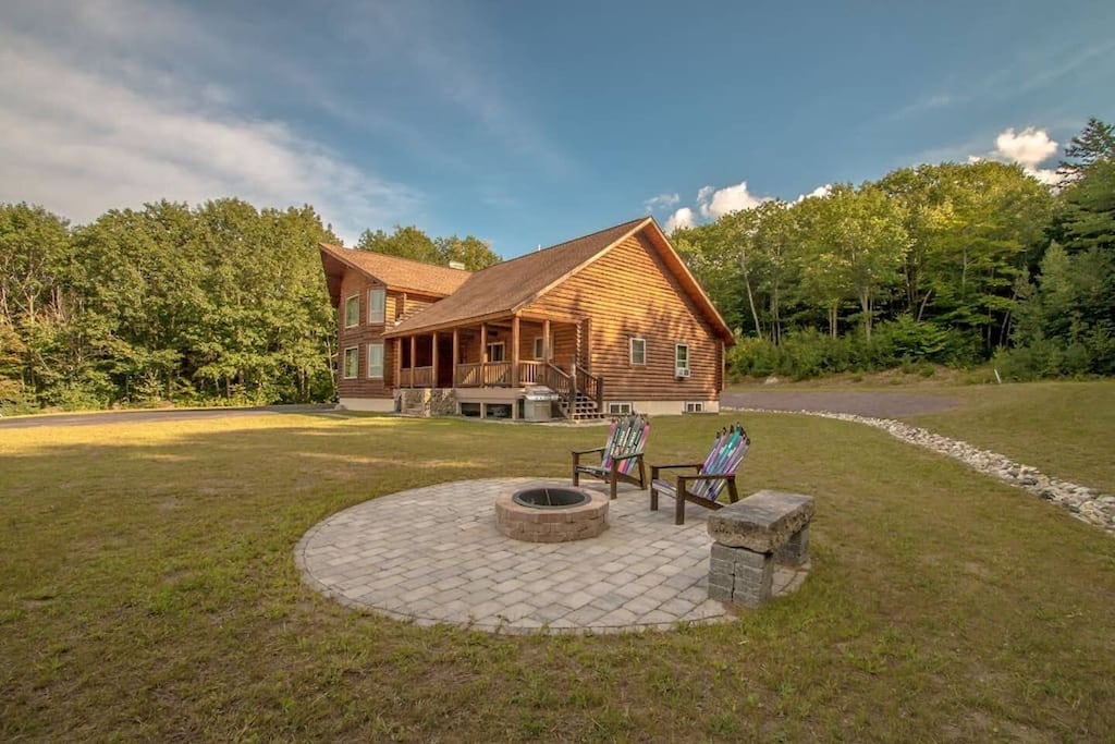 A log cabin sits surrounded by grass and trees with a fire pit and two chairs nearby