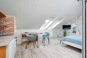  A bright attic living room with kitchen with white walls, a bed with blue bedding, and a work desk under a slanted roof. A brick wall decorates part of the room.