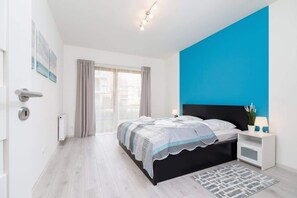 A bedroom with a double bed covered in a blue and white bedspread. The room features a bold blue accent wall and white furniture, creating a modern, fresh look.