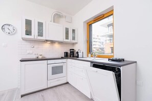 A compact kitchen with white cabinetry and modern appliances. It features a large window and minimalistic design, providing a clean and functional cooking space.