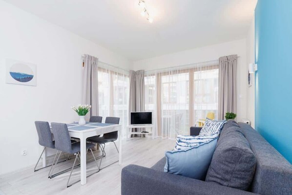 A bright living room with large windows, furnished with a white table and chairs, and a comfortable sofa. The space is airy and well-lit, with neutral decor.