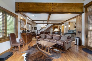 Living room with wooden beams, leather sofa, rustic coffee table, cowhide rug, and a staircase leading upstairs. The room is decorated with wooden furniture and has an open layout leading to the kitchen.