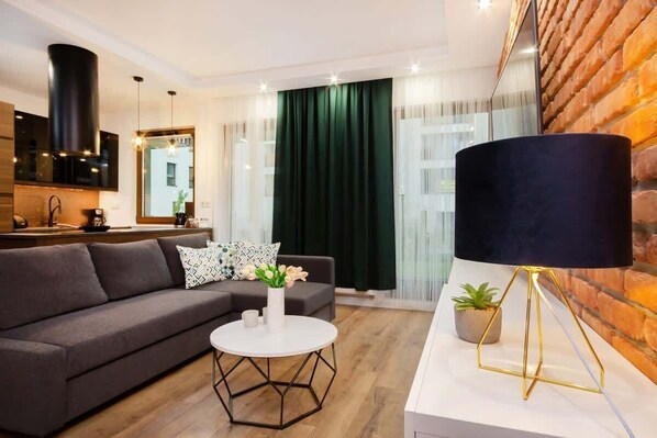 A modern living room featuring a grey sofa, a wooden coffee table, a flat-screen TV mounted on a brick accent wall, and green curtains adding a cozy touch.