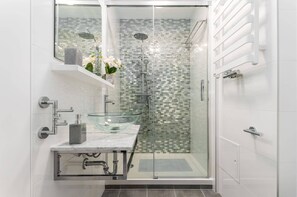 A modern bathroom with a sleek glass shower, white tiles, and a small sink, highlighted by silver accents and a bright, clean look.