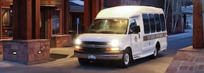 A white passenger shuttle labeled "Deer Valley Resort Skier Shuttle" is driving on a road near a stone building at dusk.