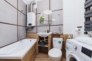 A small bathroom with a bathtub, toilet, sink, and washing machine. Tiled walls and a shelf above the toilet add functionality.