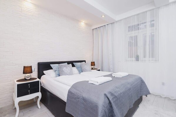 A bright bedroom featuring a large, comfortable bed covered with gray throws. Soft lighting and white curtains add elegance and coziness to the space.