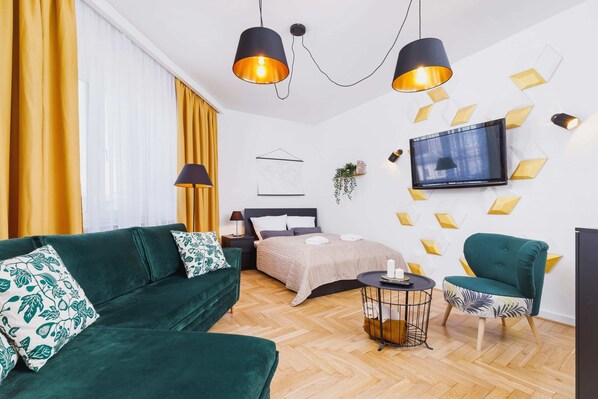 A bright interior featuring a large green sofa with decorative cushions and a modern coffee table. In the background, there’s a wall-mounted TV and yellow curtains adding a vibrant touch.
