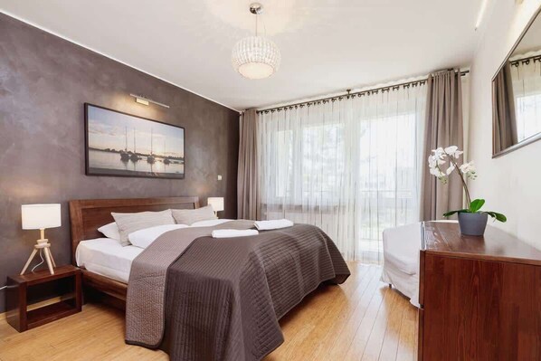  A bedroom with a large bed featuring dark bedding, wooden floors, and natural light coming through a window with sheer curtains. The room feels cozy and warm.