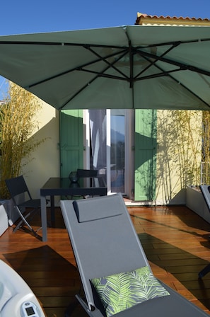 La terrasse avec table, grand parasol, chaises longues et jacuzzi