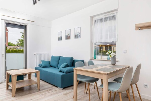 A cozy living room featuring a modern blue sofa and a wooden coffee table, set against a bright, airy background with large windows and simple wall art.