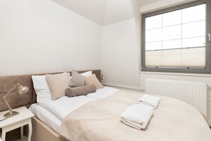 A cozy bedroom featuring a double bed adorned with light-colored linens. A large window fills the room with soft natural light.