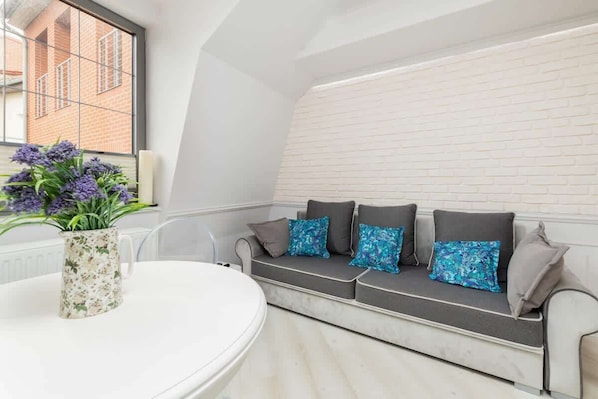 A cozy living room featuring a gray sofa with vibrant turquoise pillows and a round white dining table. The space has soft lighting and a modern yet inviting design.