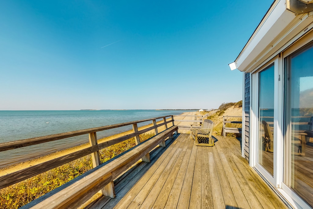 A long, narrow wooden balcony looks out from an oceanfront Cape Cod vacation rental on a sunny day