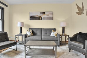 A grey upholstered couch with end tables and lamps on both ends, grey coffee table and two club chairs.