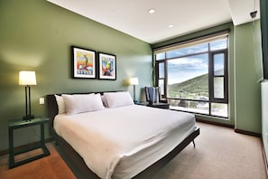 Main bedroom with luxurious king bed, white duvets and linens, black leather accent chair and bright window wall