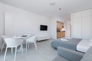 A spacious and minimalistic living room with a gray bed, armchair, coffee table, and a large window allowing in natural light.